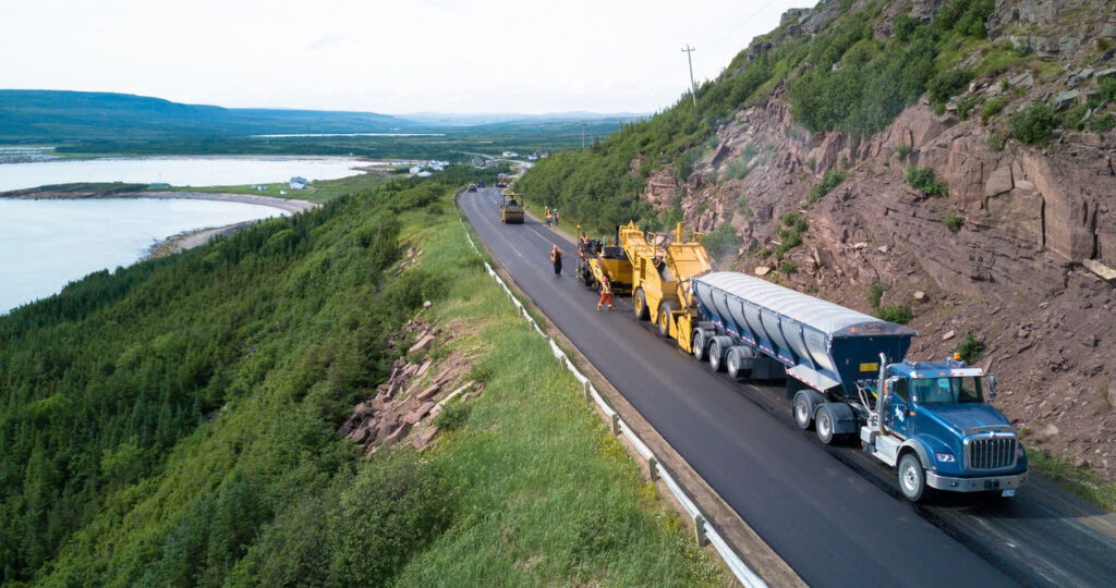 A tarp-covered transport truck and road paving crew on a coastal Newfoundland highway